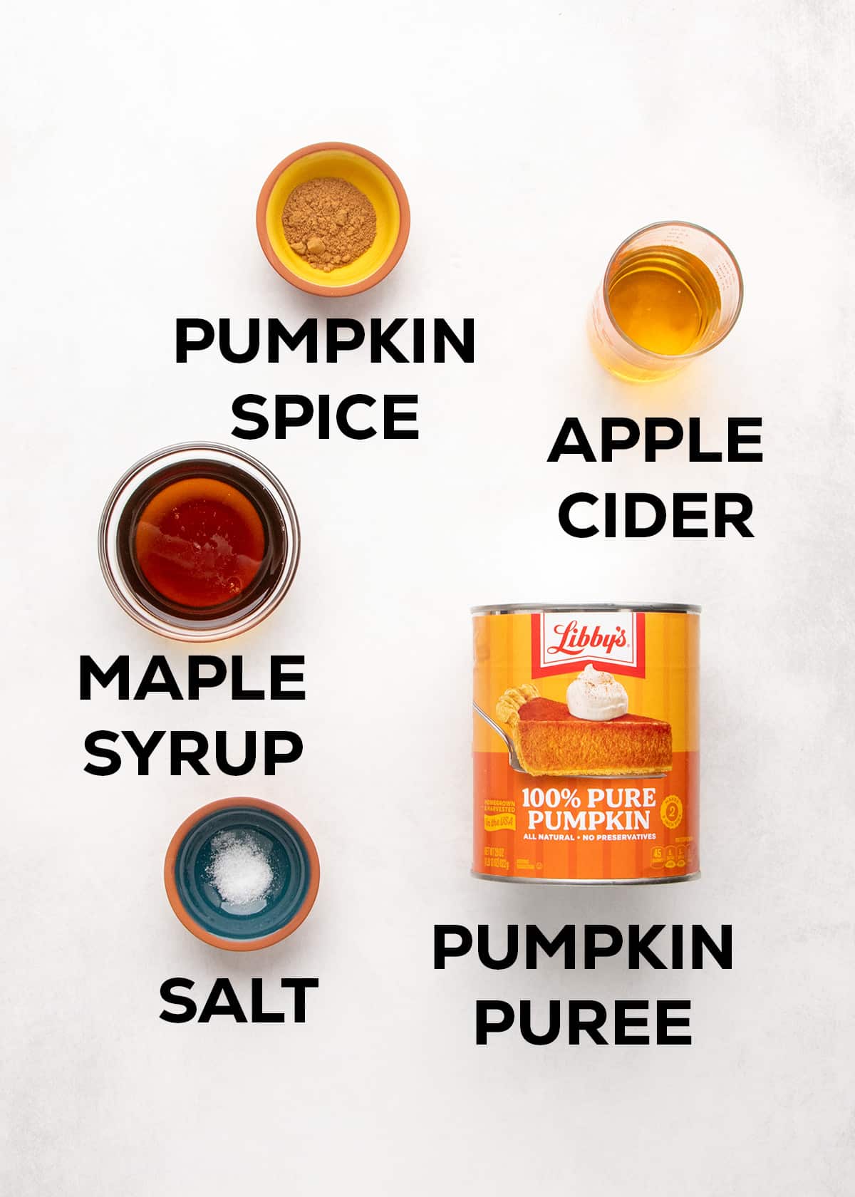 Bowls and containers labeled pumpkin spice, apple cider, maple syrup, salt, and pumpkin puree sit on a white surface.