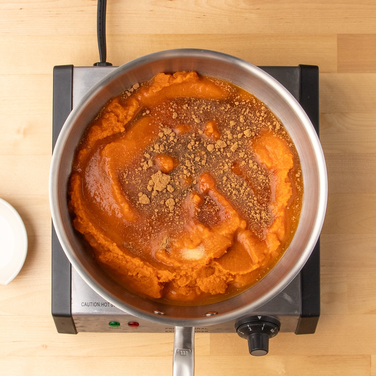 Pumpkin puree, liquid, and spices sit together but uncombined in a stainless steel pan on an electric burner.